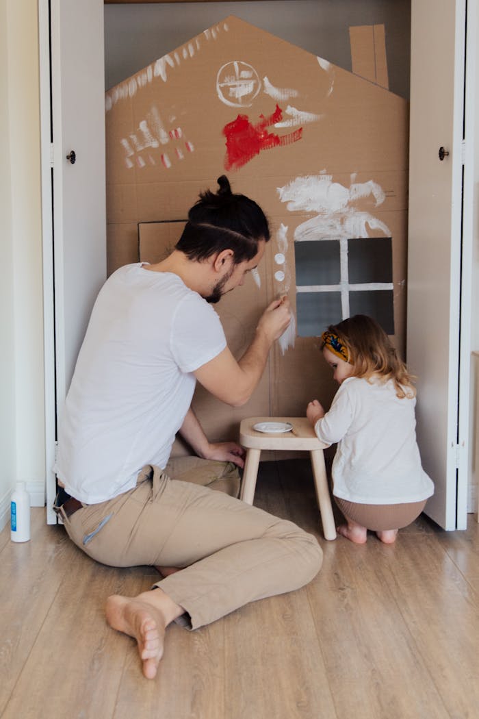 gallery-4 Father and daughter painting a cardboard house indoors, enjoying quality time together.