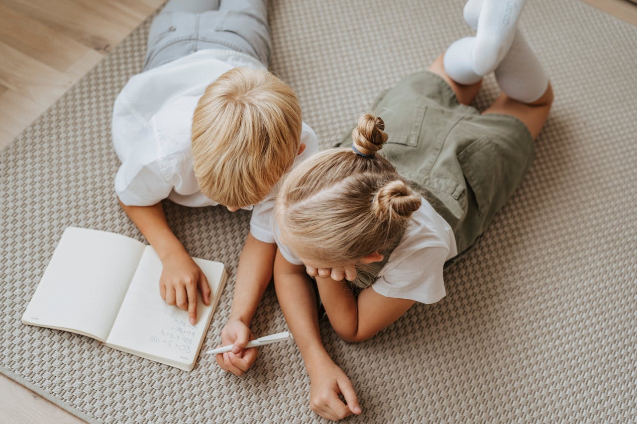 our-services-3 Two young children study together on the floor, concentrating on a notebook in a warm indoor setting.